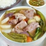 Sinigang na Baboy in a white bowl with a plate of steamed rice and a small bowl of patis in the background.