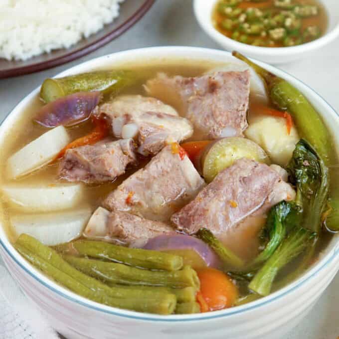Sinigang na Baboy in a white bowl with a plate of steamed rice and a small bowl of patis in the background.