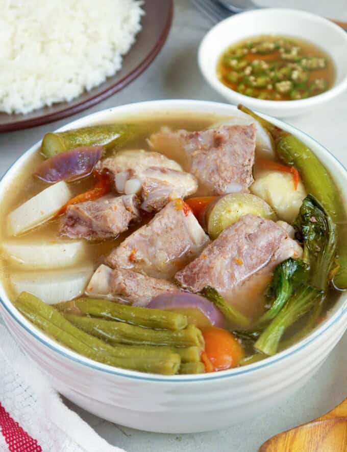 Sinigang na Baboy in a white bowl with a plate of steamed rice and a small bowl of patis in the background.