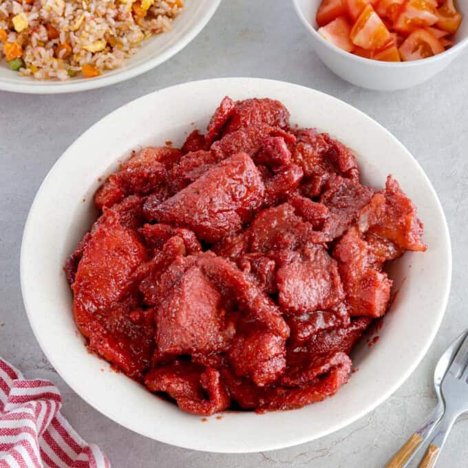 pork tocino in a white serving bowl with fried rice and sliced tomatoes in the background.