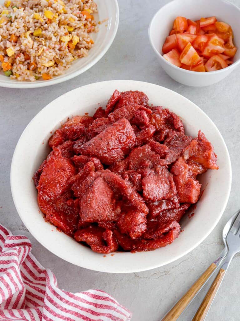 pork tocino in a white serving bowl with fried rice and sliced tomatoes in the background.