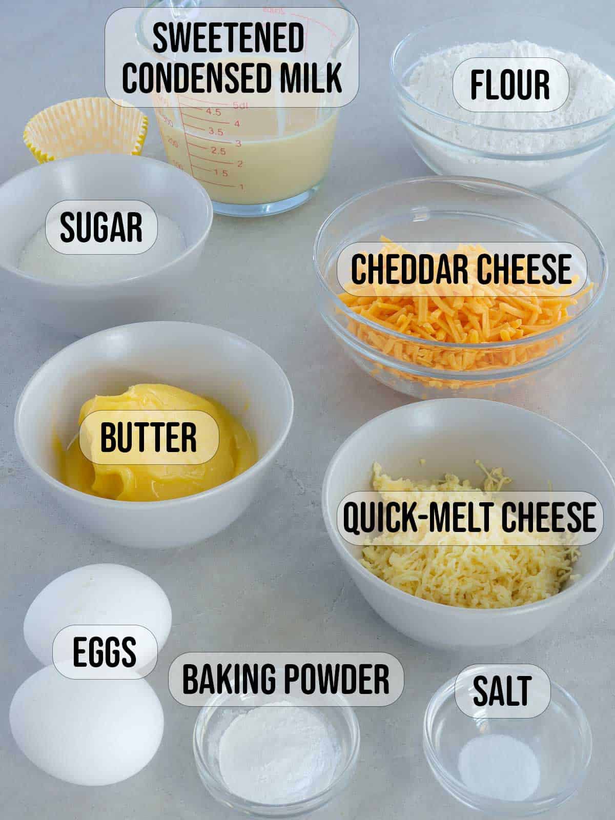 flour, condensed milk, sugar, eggs, butter, baking powder, salt, shredded cheddar, shredded quick melt cheese in bowls.