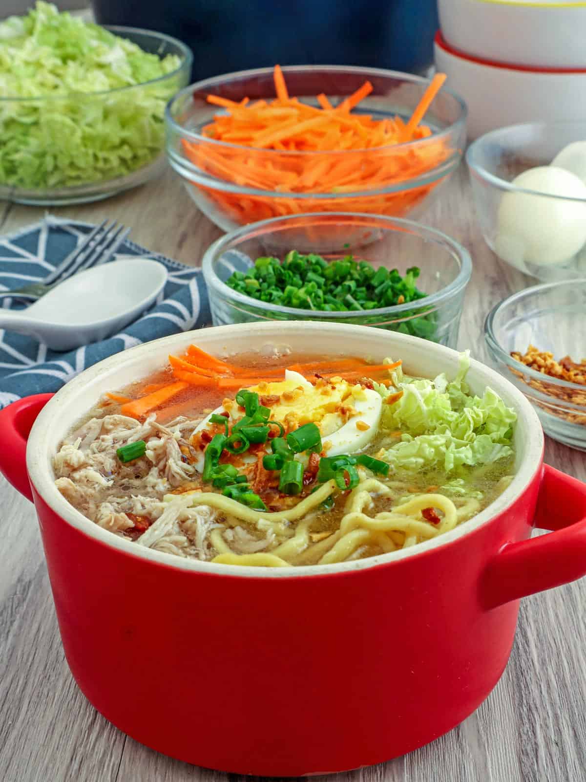 chicken mami in a red bowl with vegetables, hardboiled eggs, and fried garlic in the background.