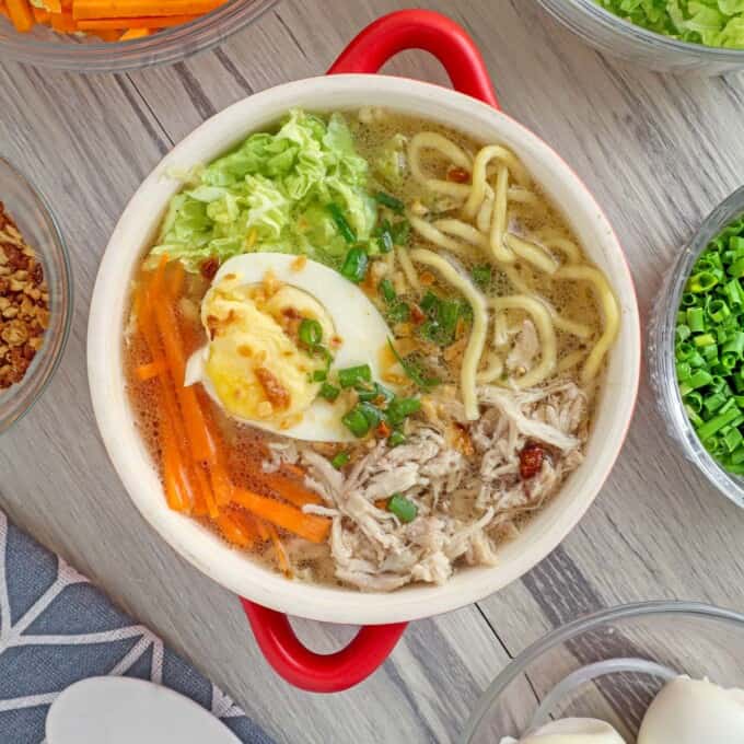 Chicken Mami in a red bowl with assorted veggies and egg toppings in the background.