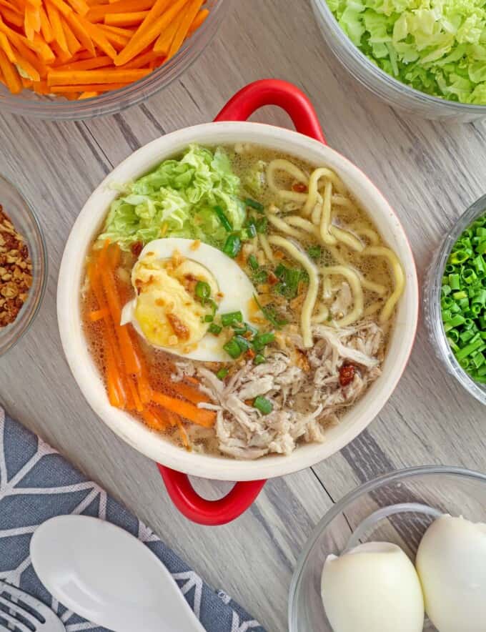 Chicken Mami in a red bowl with assorted veggies and egg toppings in the background.