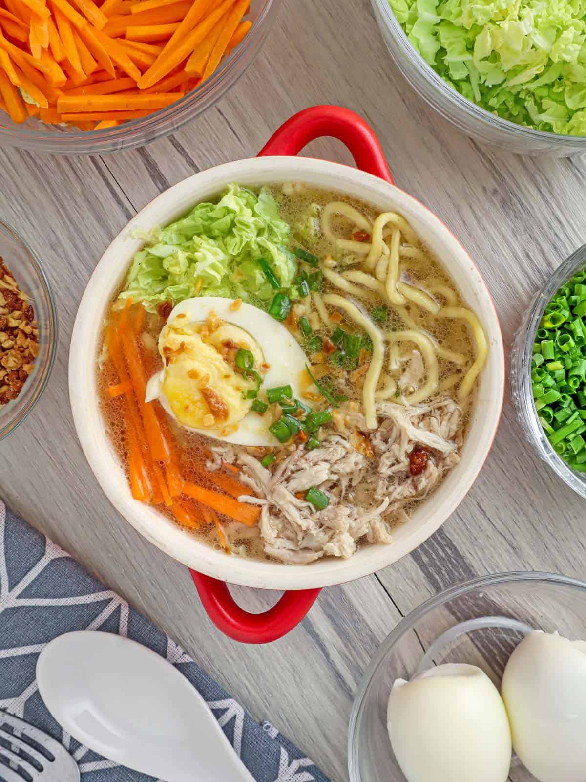 Chicken Mami in a red bowl with assorted veggies and egg toppings in the background.