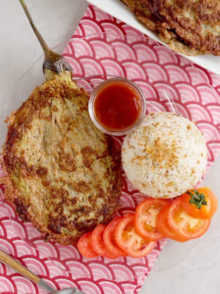 Tortang Talong on a clear glass plate with steamed rice and sliced tomatoes on the side
