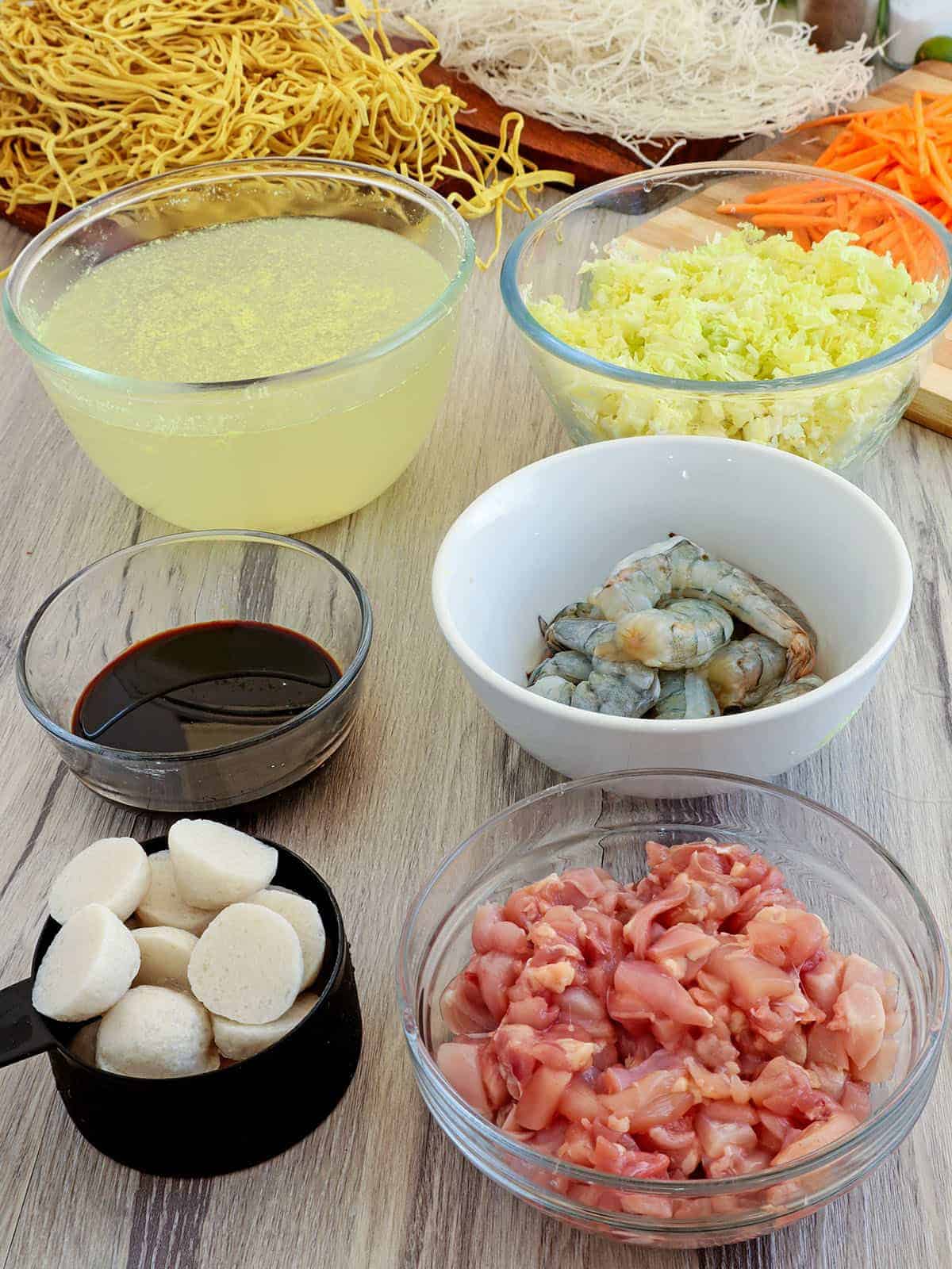 fresh egg noodles, rice stick noodles, sliced chicken, fish balls, peeled shrimp, chicken broth, shredded cabbage, julienned carrots, kecap manis in individual bowls