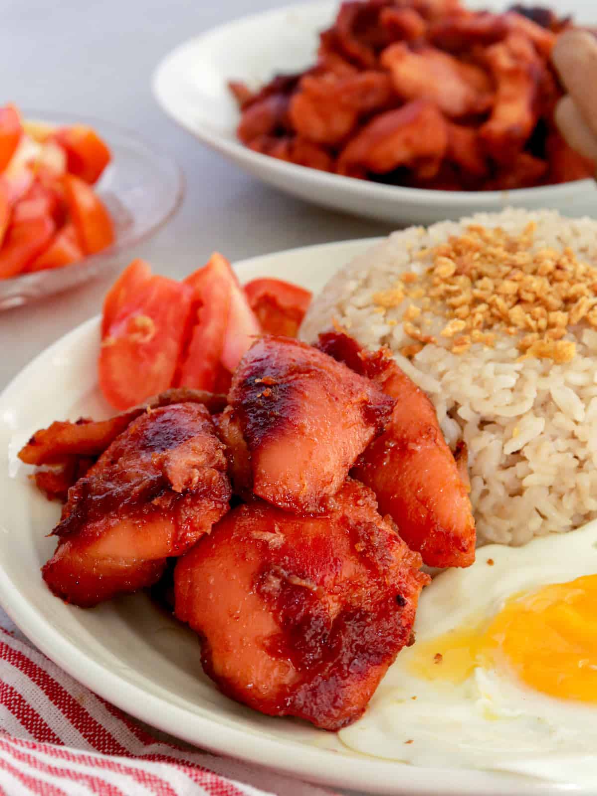 chicken tocino on a white plate with a side of fried egg, garlic rice, and sliced tomatoes.