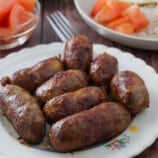 cooked longganisa on a white plate with chopped tomatoes, fried rice, and fried eggs in the background.