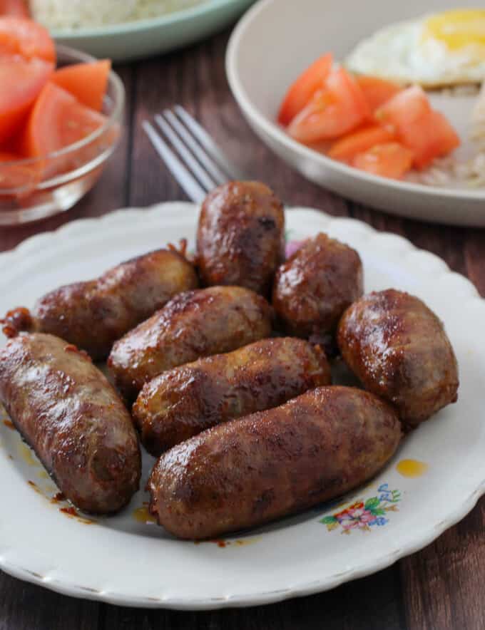 cooked longganisa on a white plate with chopped tomatoes, fried rice, and fried eggs in the background.