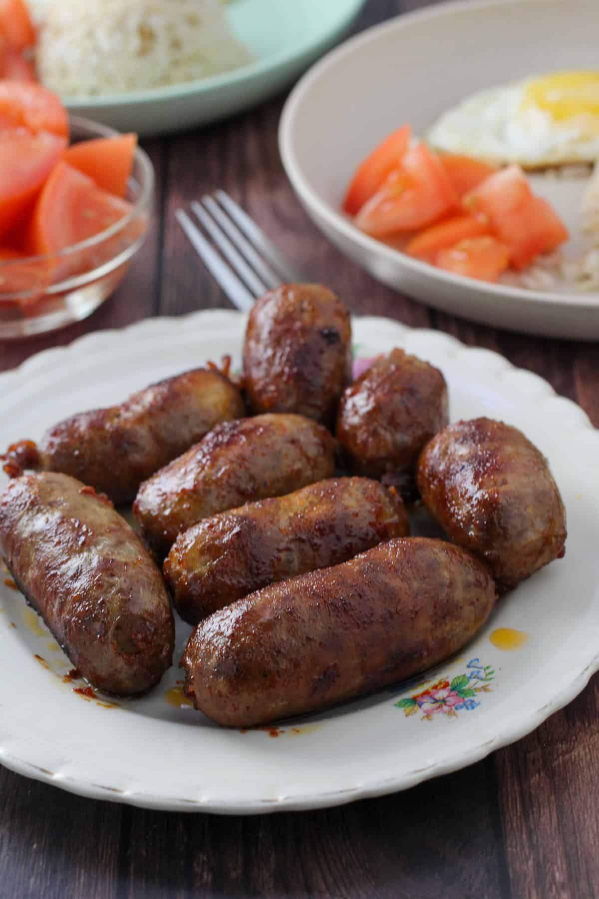 cooked longganisa on a white plate with chopped tomatoes, fried rice, and fried eggs in the background.