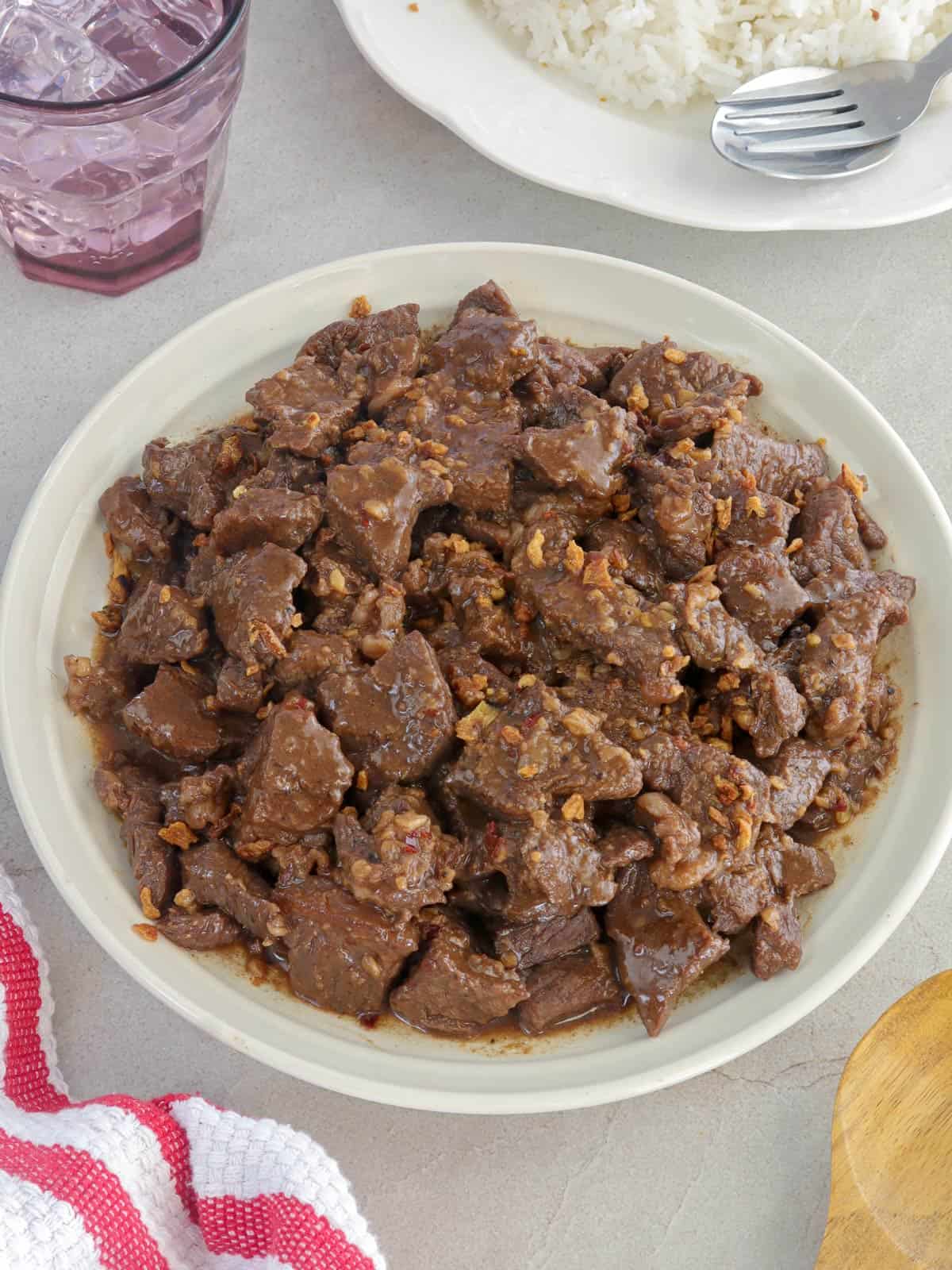 Beef Salpicao on a serving plate with steamed rice and glass of water in the background.