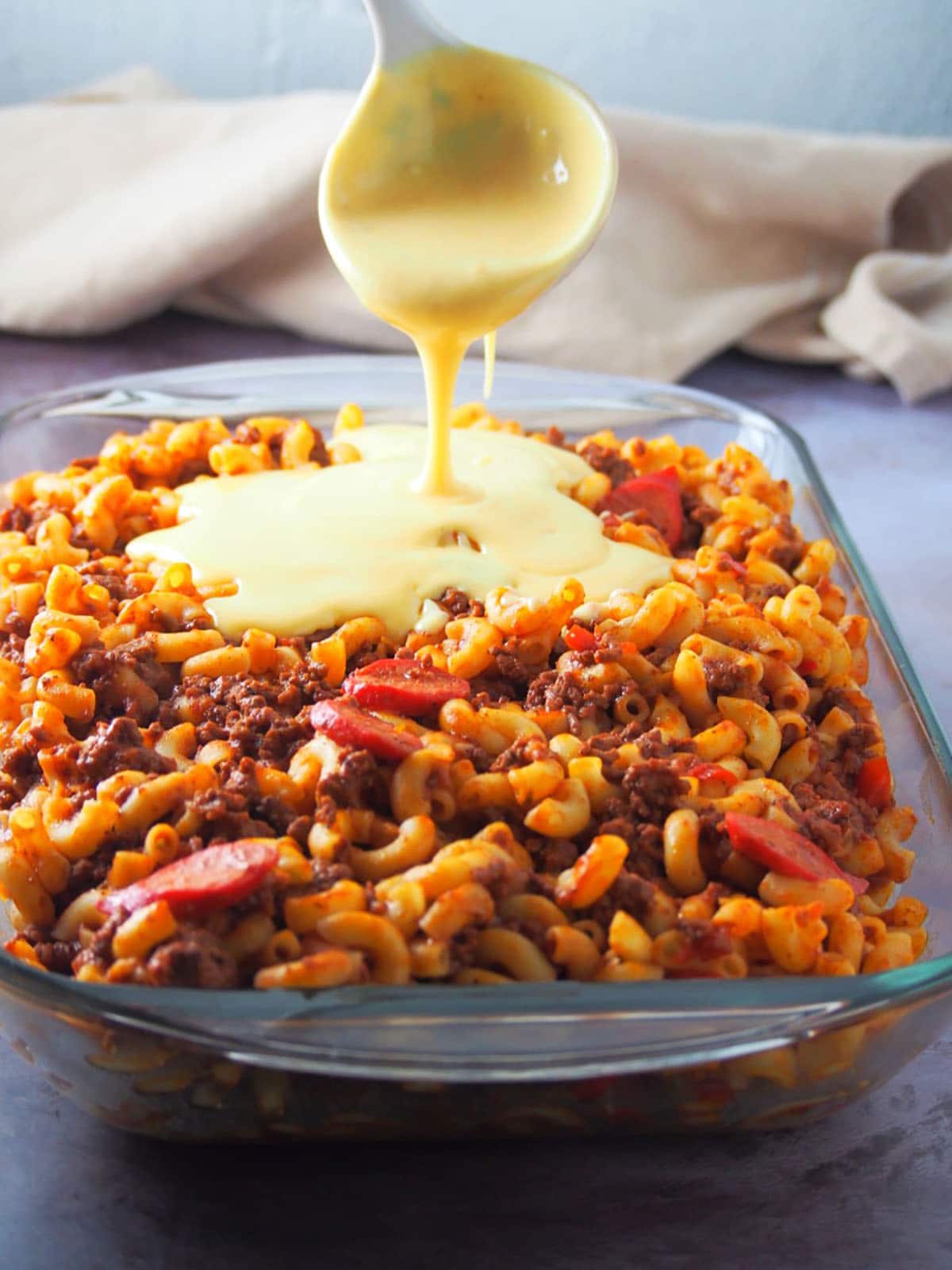 pouring cheese sauce topping on beef macaroni in a baking dish