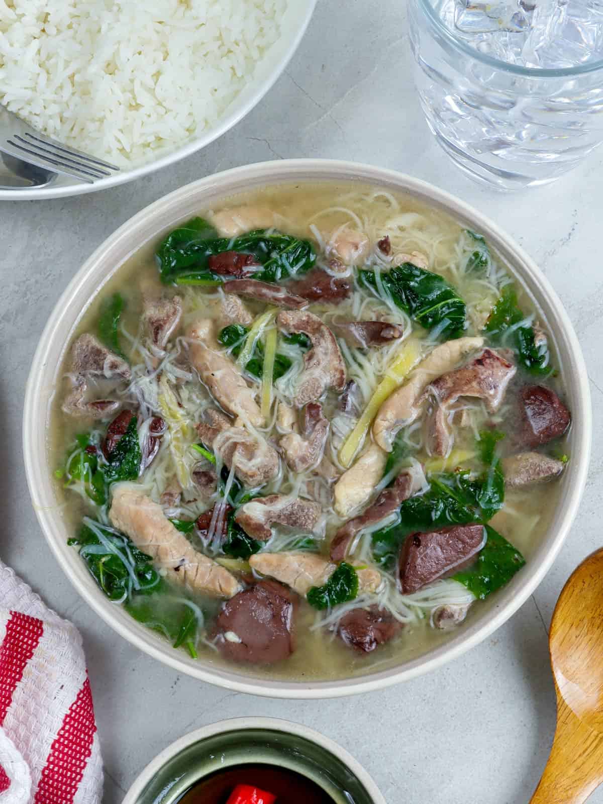 Batchoy Tagalog in a white bowl with a glass of water and a plate of steamed rice in the background.