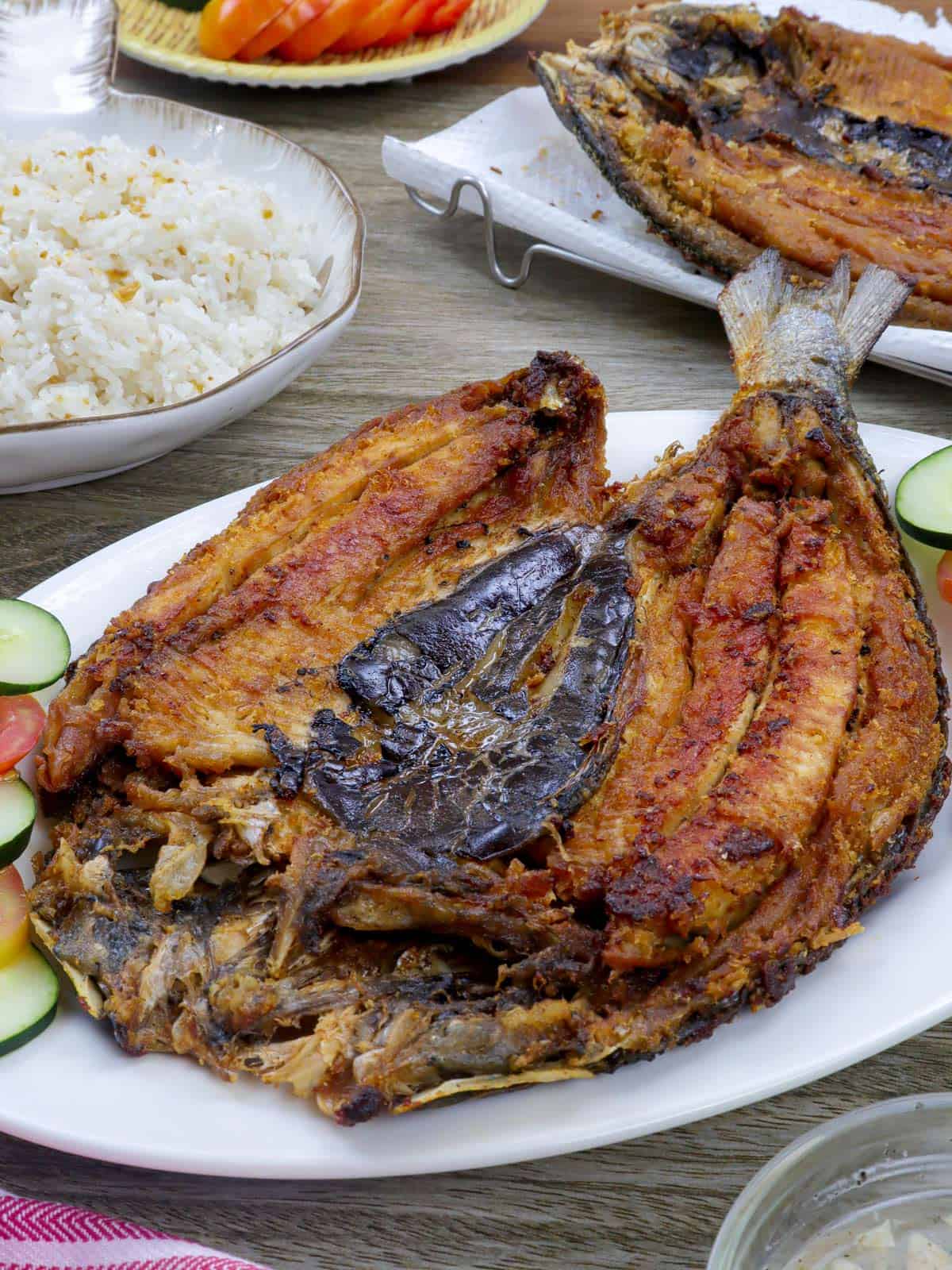 fried butterflied milkfish on a white plate with steamed rice and sliced tomatoes and cucumber on the side and plate of fried rice in the background.