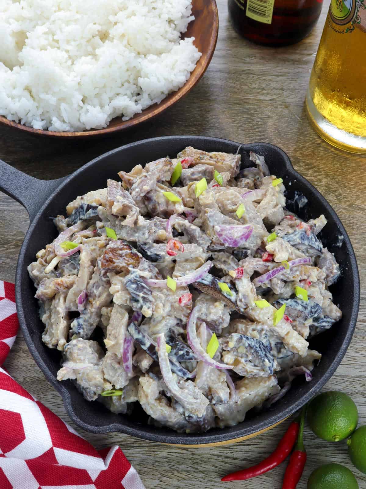 Dinakdakan in a cast iron skillet with a bowl of steamed rice on the side and a glass of beer