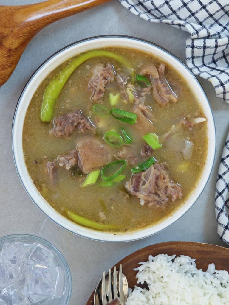 Sinampalukang Kambing in a white serving bowl with a plate of steamed rice on the side