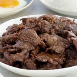 beef tapa on a white serving dish with plates of steamed rice and fried eggs in the background.
