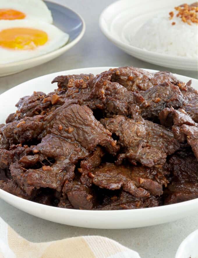 beef tapa on a white serving dish with plates of steamed rice and fried eggs in the background.