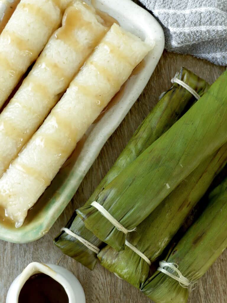 Suman Malagkit wrapped in banana leaves on a serving platter with a bowl of caramel coconut sauce on the side