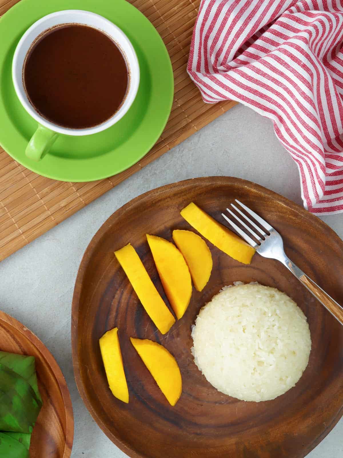 puto maya with sliced mangoes on a wooden plate and a cup of sikwate