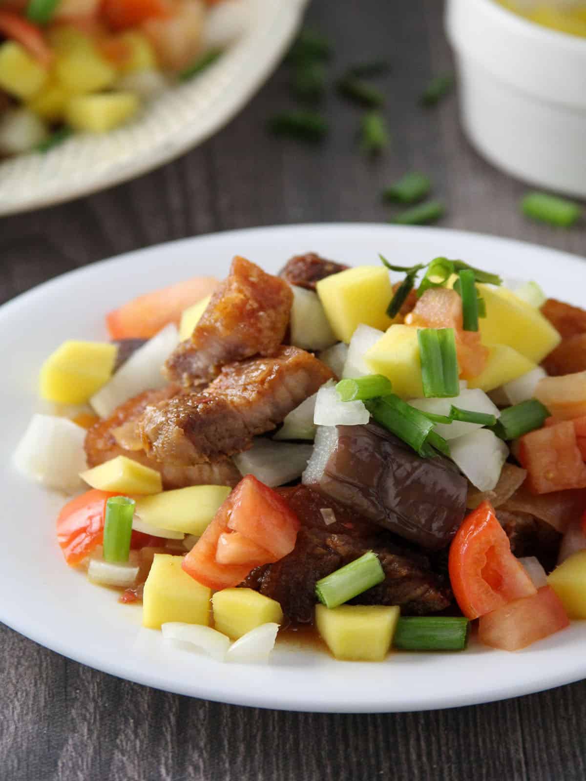 Crispy Pork Binagoongan topped with chopped mangoes, tomatoes, steamed eggplants, and green onions on a plate