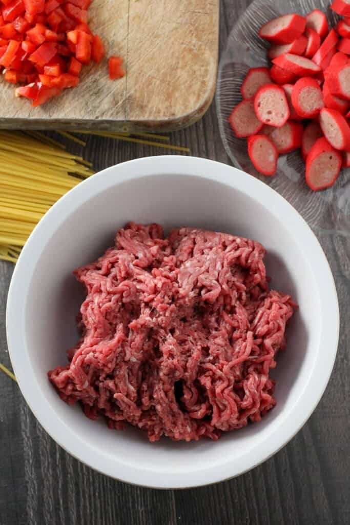 uncooked ground beef in a white bowl, dry spaghetti, sliced hotdogs on a clear plate, and chopped red bell peppers on a cutting board