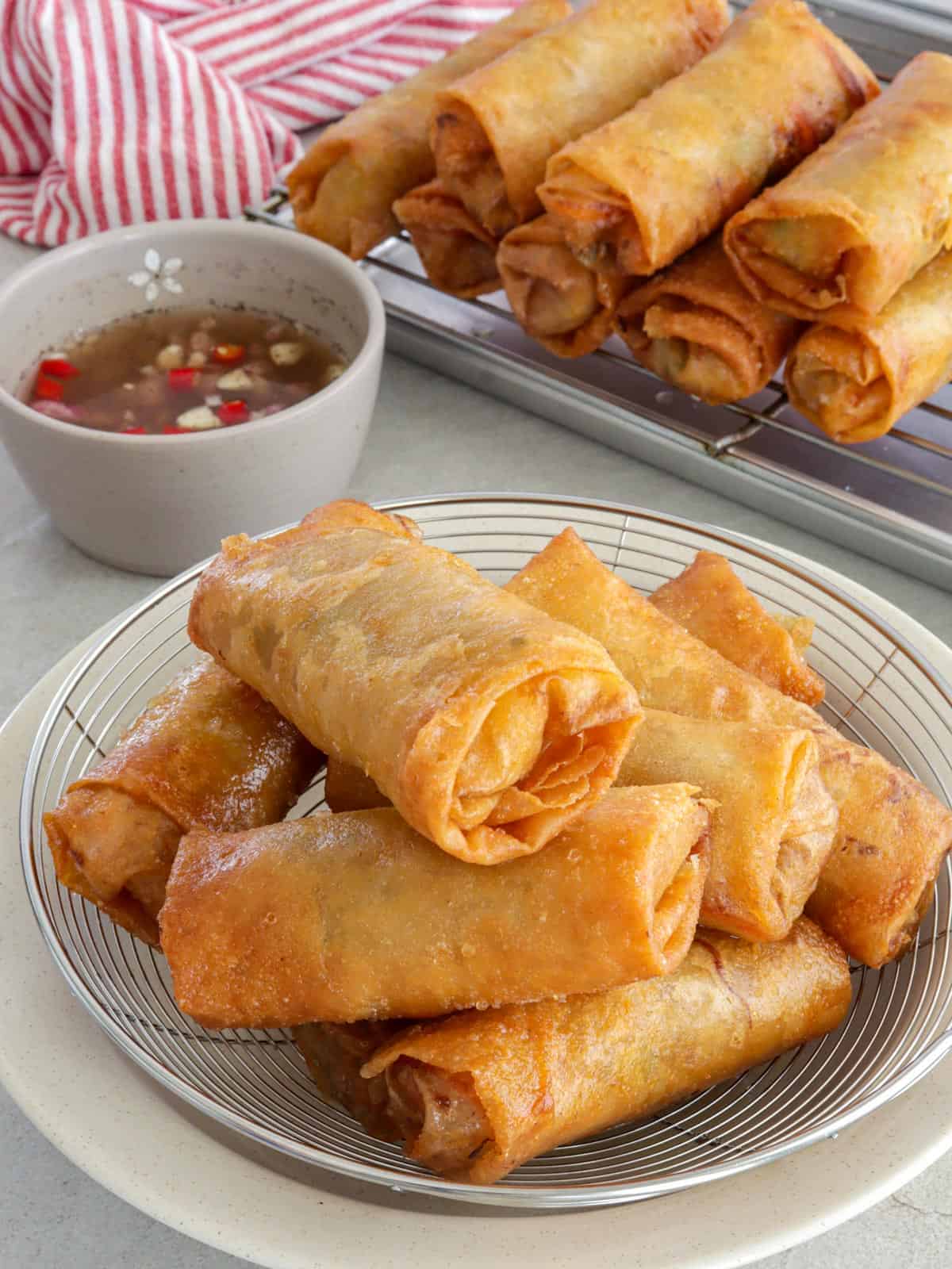 crispy Lumpiang Togue on a white serving plate with a small bowl of spiced vinegar and more fried lumpia on a wire rack in the background.