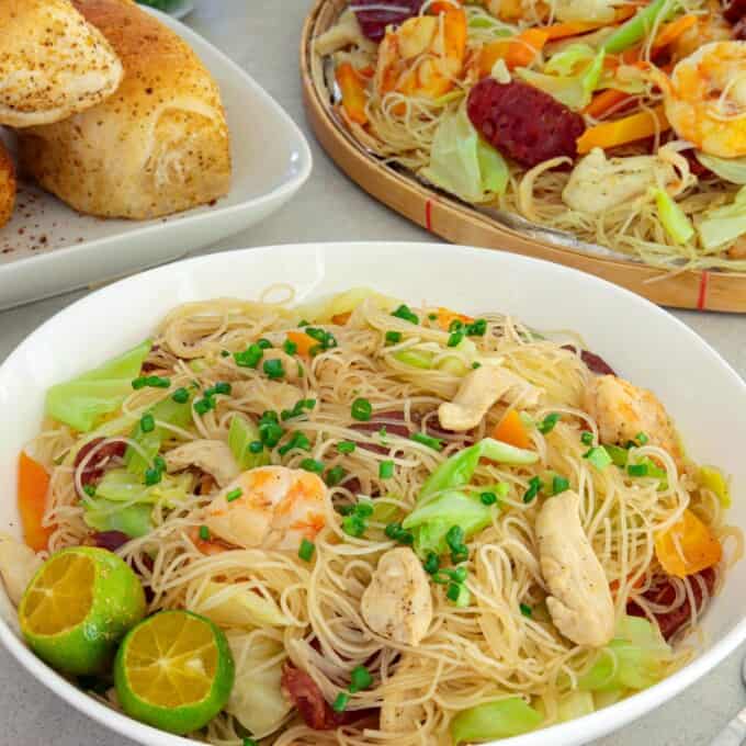 bihon guisado in a white serving bowl with pandesal and a bilao of pancit on the side.