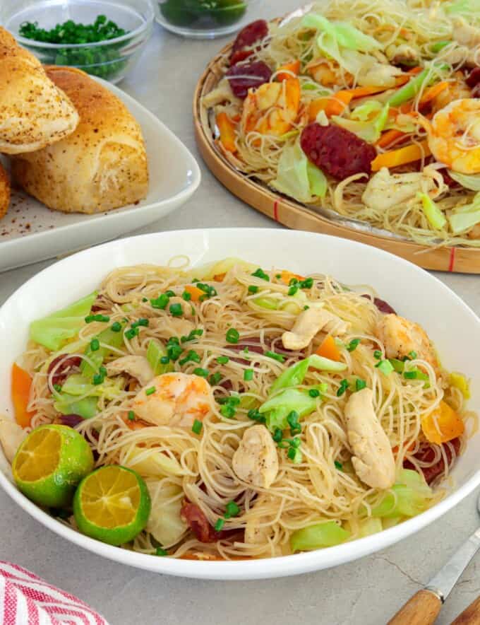 bihon guisado in a white serving bowl with pandesal and a bilao of pancit on the side.