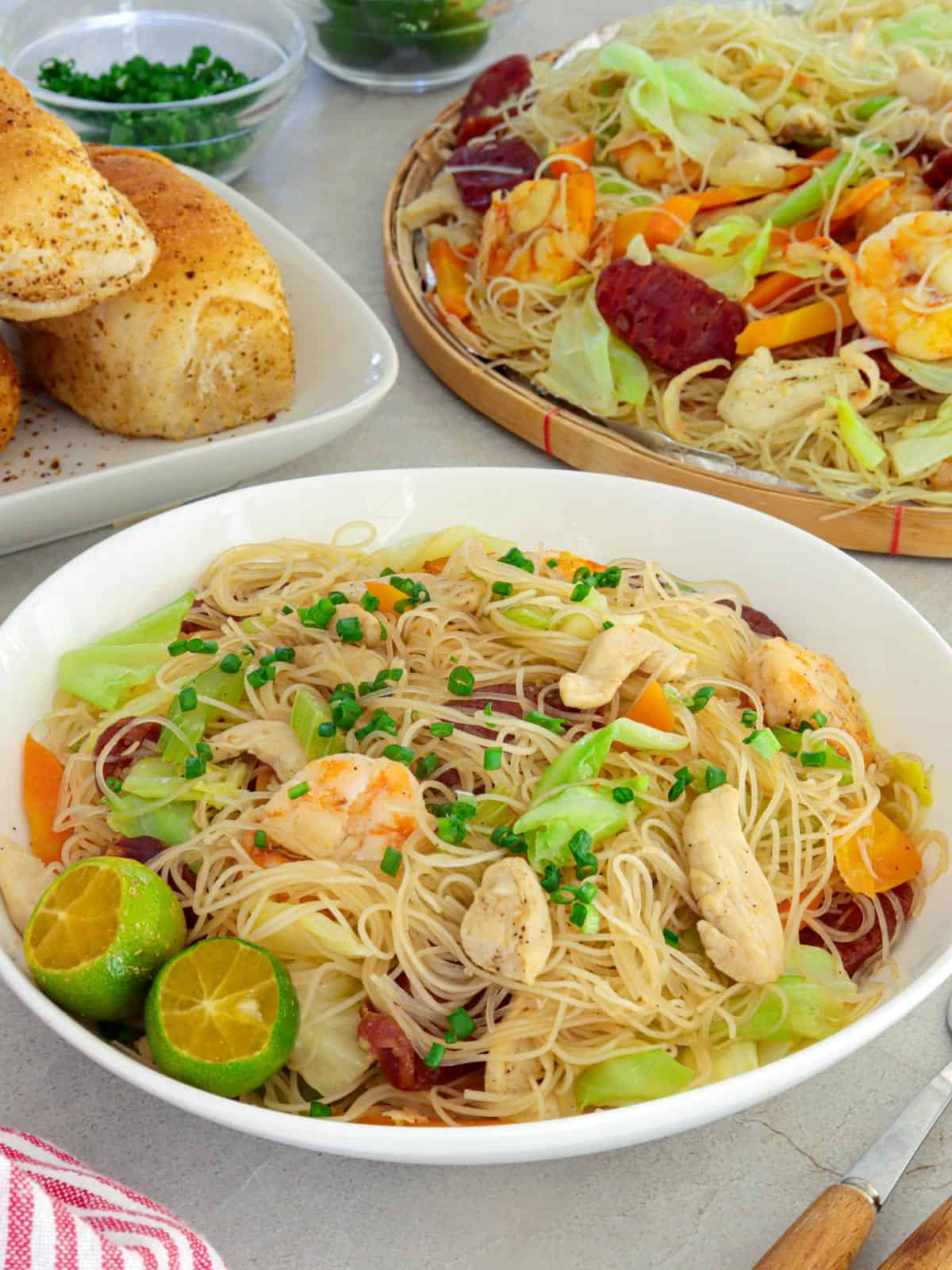 bihon guisado in a white serving bowl with pandesal and a bilao of pancit on the side.