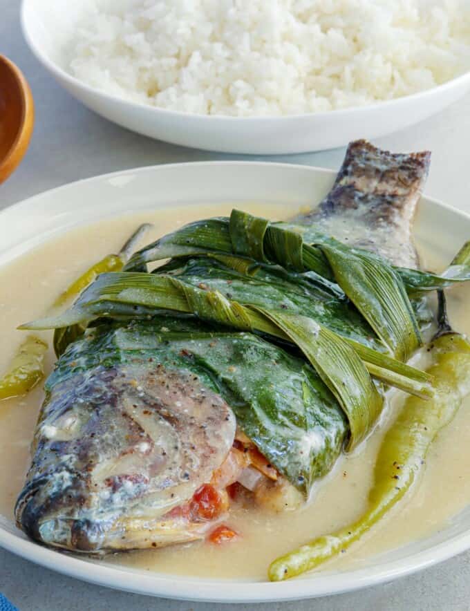 Sinanglay na Tilapia on white serving plate with a plate of steamed rice in the background.
