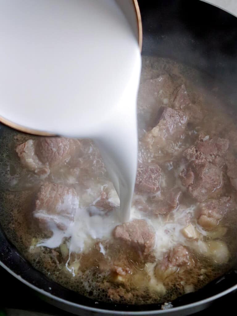 pouring coconut milk into a pan of beef cubes and broth