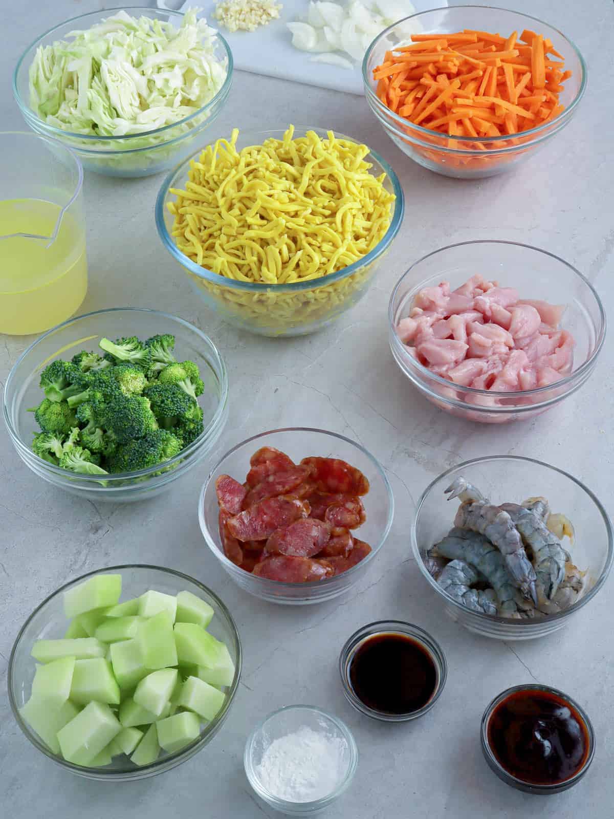 lomi noodles, sliced chicken breasts, broccoli florets, peeled shrimp, julienned carrots, chopped onions, sliced chayote, sliced Chinese sausage in individual bowls