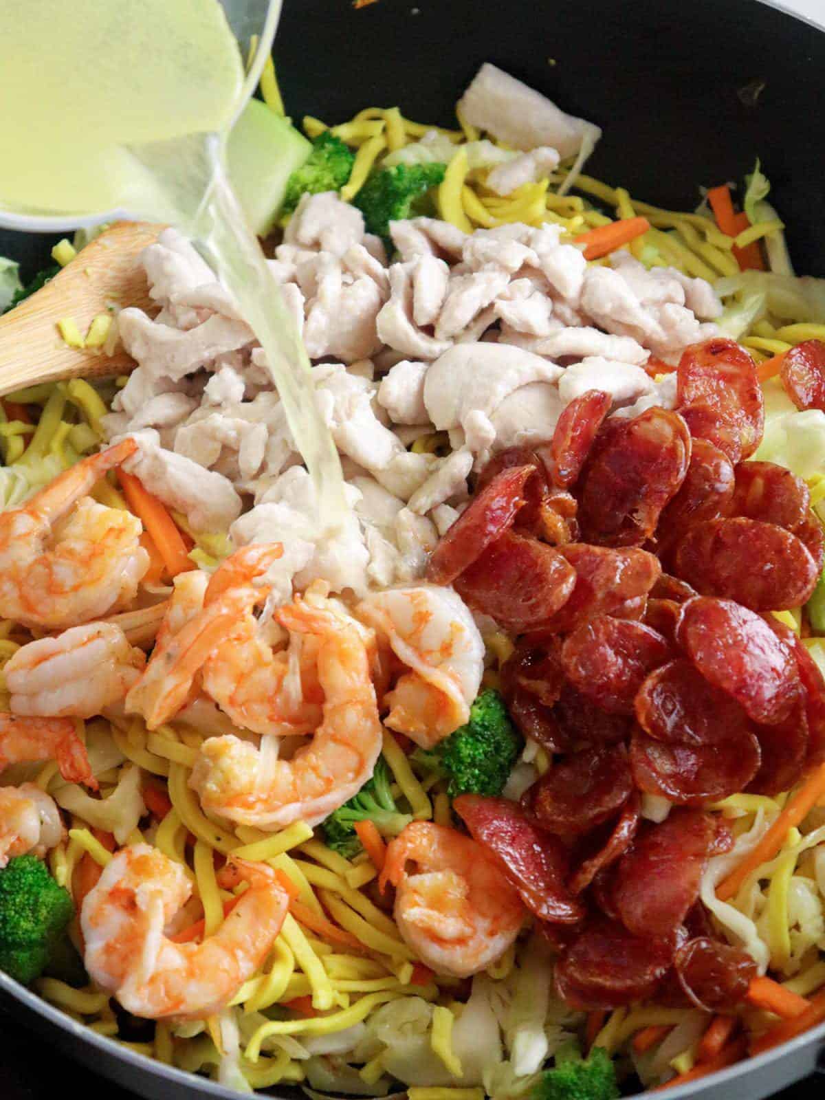 adding chicken broth to shrimp, Chinese sausages, chicken, assorted veggies and noodles in a skillet