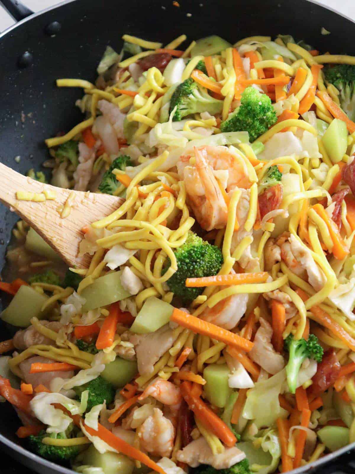 stirring pancit lomi guisado in a skillet with a wooden spoon