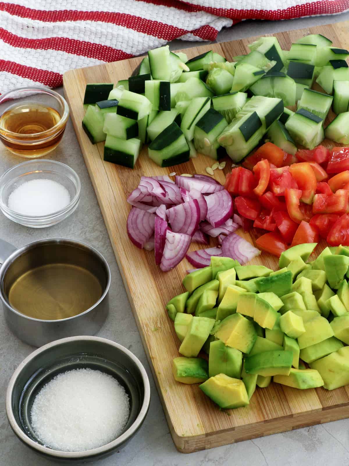 diced cucumbers, red onions, tomatoes, cucumbers on a wooden board with bowls of sugar, salt, pepper, and sesame oil on the side