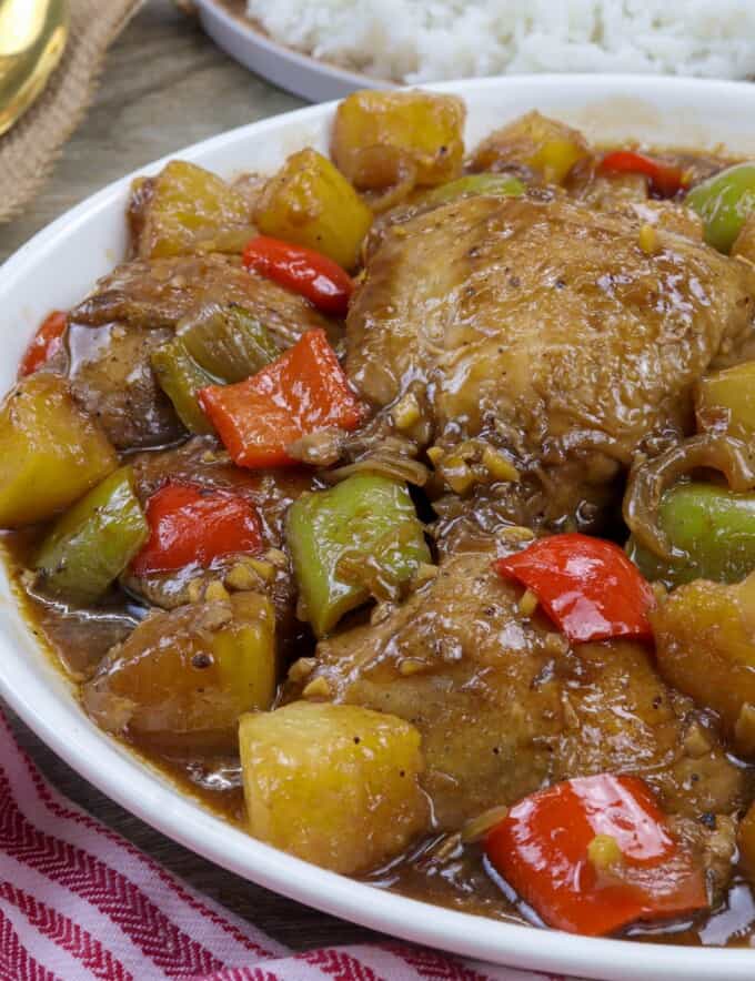 Hawaiian Chicken in a white serving bowl with a plate of steamed rice in the background.