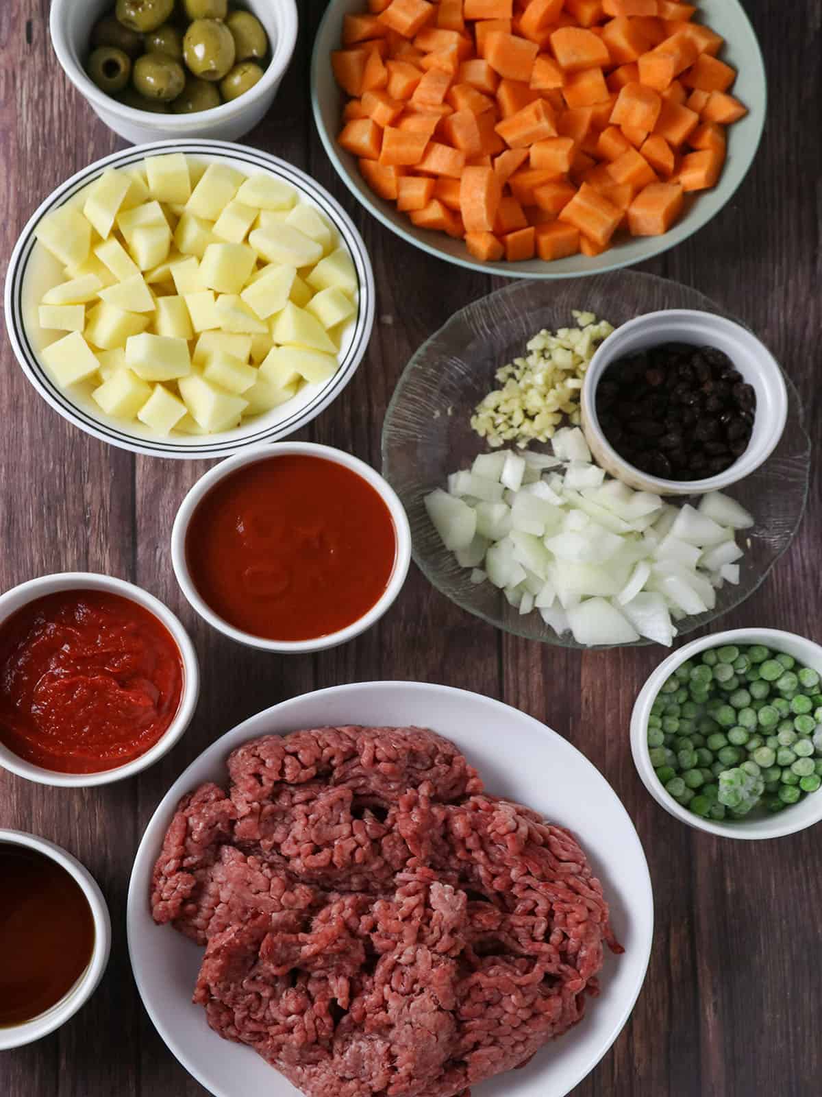 ground beef, diced potatoes, carrots, green peas, olives, raisins, tomato sauce, onions, garlic in bowls