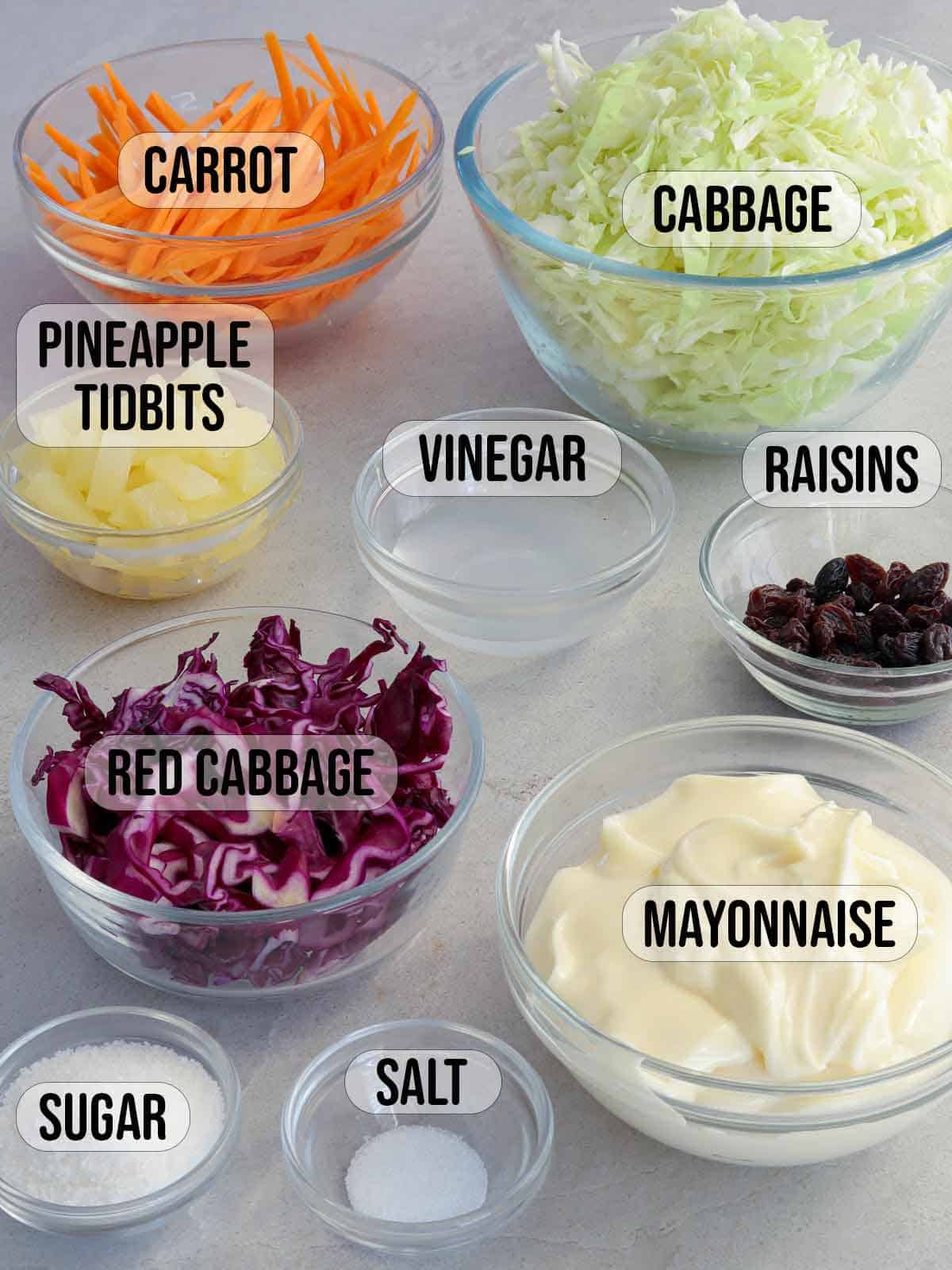 shredded cabbage, shredded red cabbage, shredded carrtos, pineapple tidbits, mayonnaise, vinegar, raisins, sugar, salt in bowls.