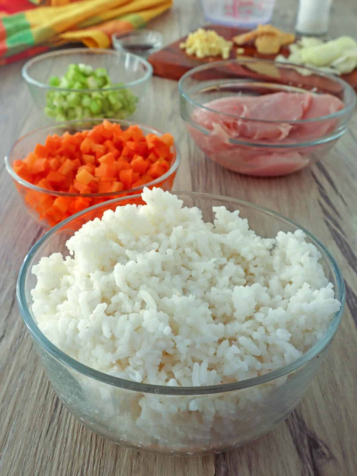 steamed rice, diced carrots, diced celery, uncooked chicken breast, chopped onions in bowls