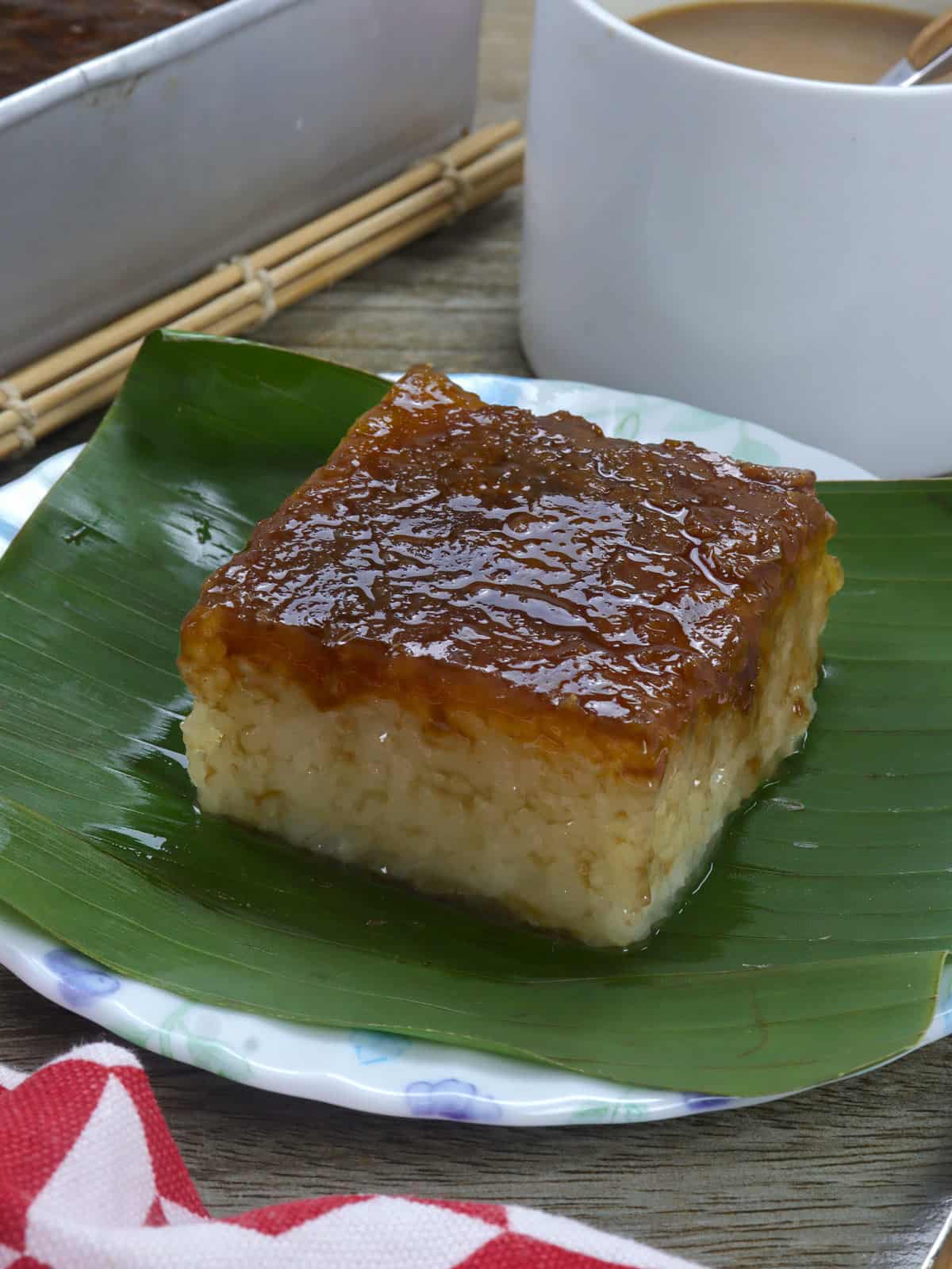 slice of bibingkang malagkit on a banana-lined white serving plate with cup of coffee in the bakcground.