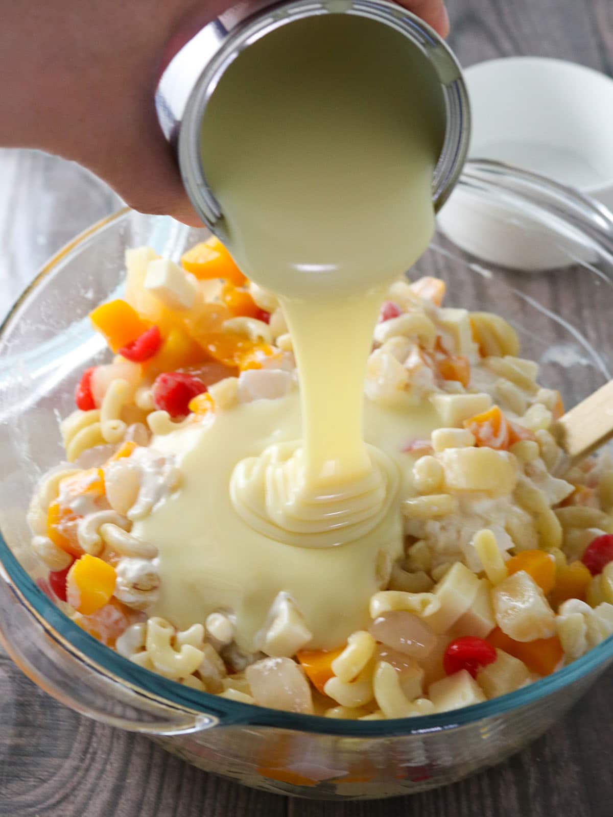 pouring condensed milk on fruit salad with macaroni in a glass bowl