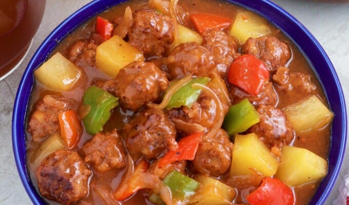 Sweet and Sour Meatballs with bell peppers and pineapples in a a blue bowl with a plate of steamed rice on the side