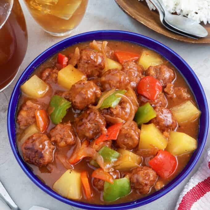 Sweet and Sour Meatballs with bell peppers and pineapples in a a blue bowl with a plate of steamed rice on the side