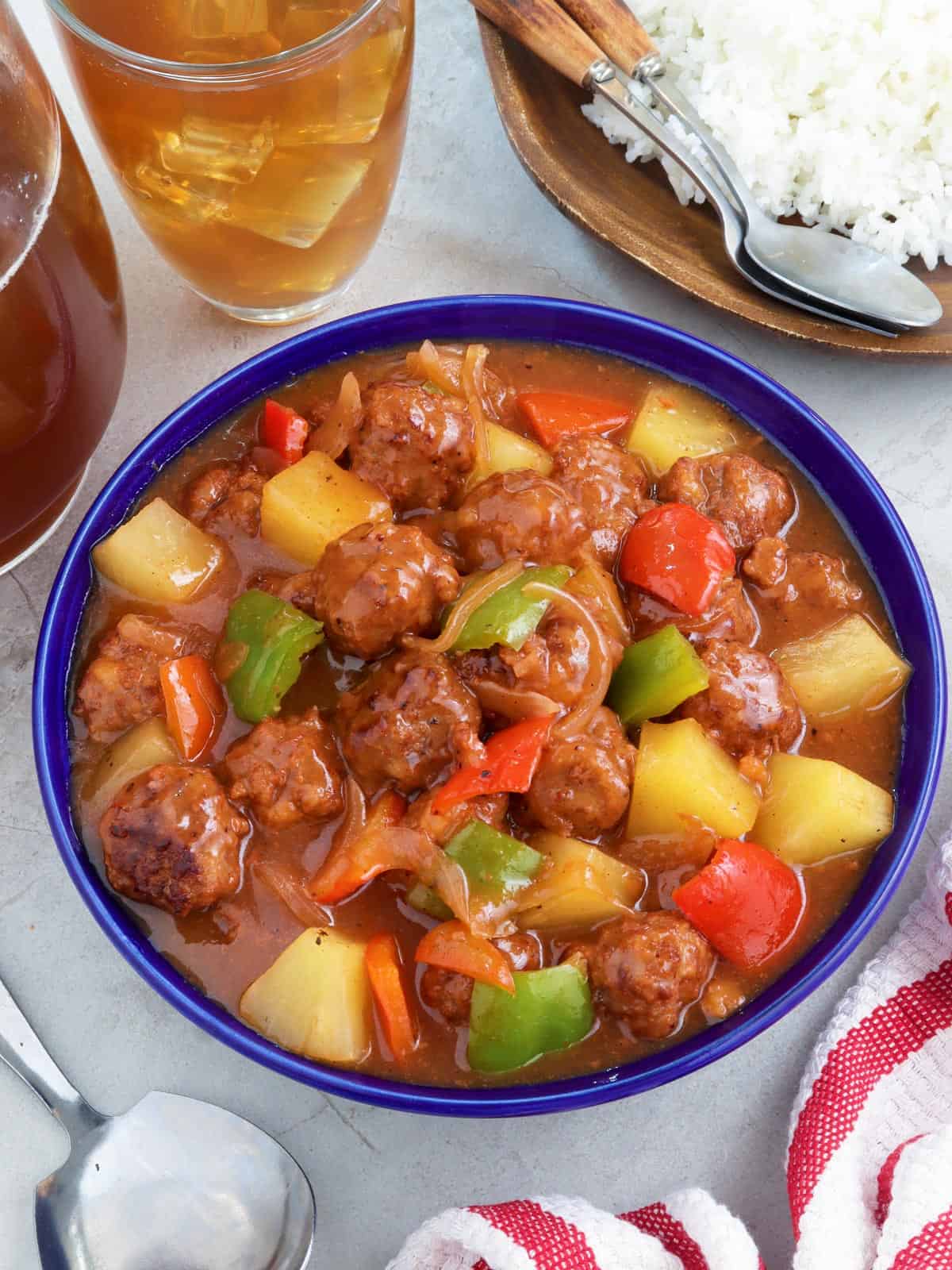 Sweet and Sour Meatballs with bell peppers and pineapples in a a blue bowl with a plate of steamed rice on the side