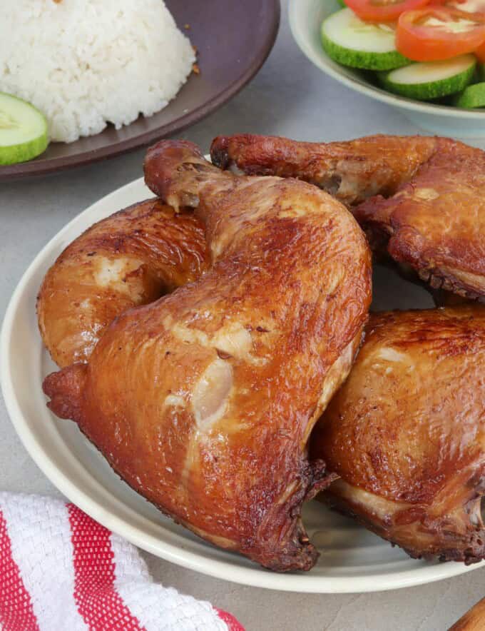 soy sauce chicken on a white platter with rice and sliced cucumbers and tomatoes on the side.