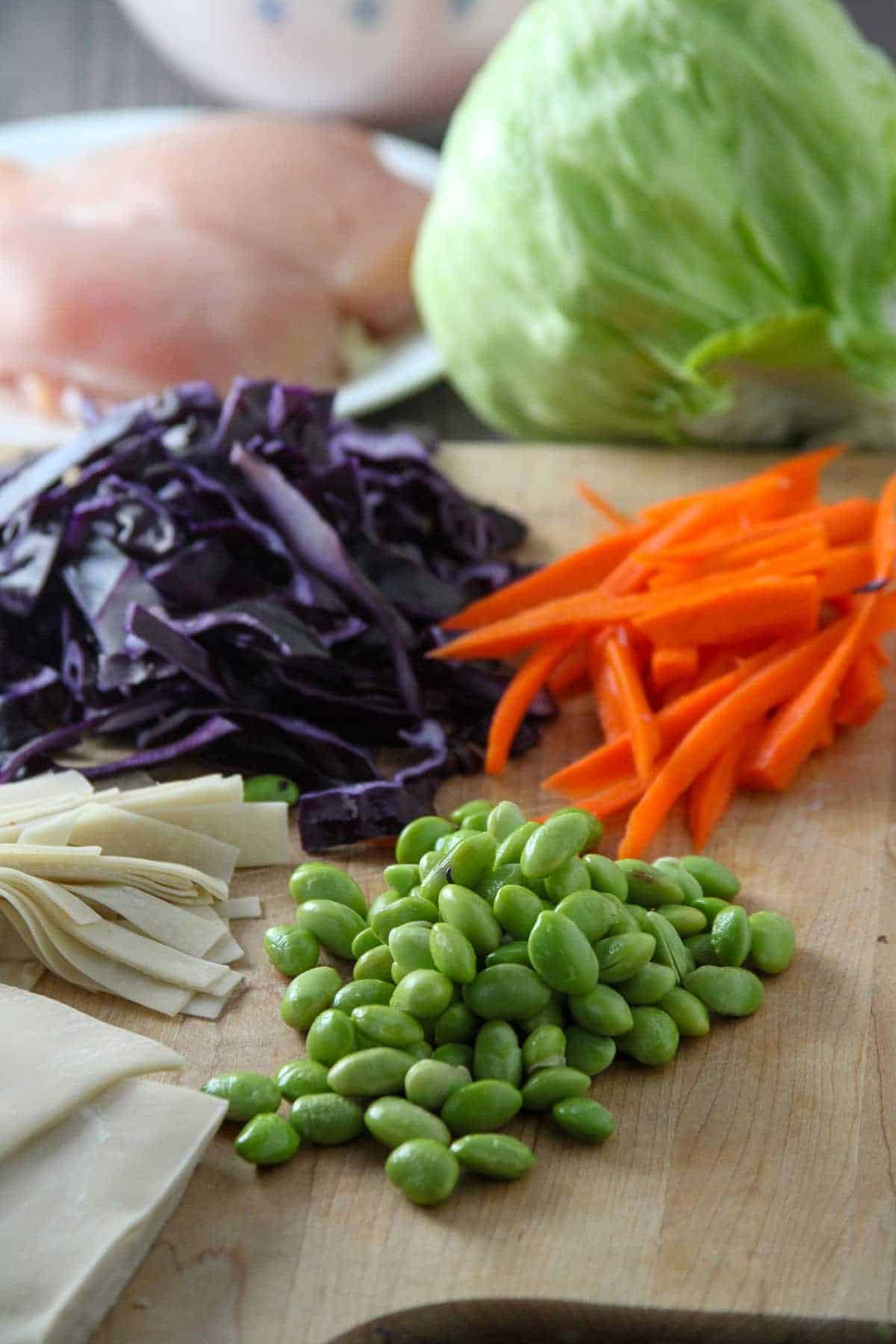 edamame, cut wonton kins, julienned carrots, shredded red cabbage, iceberg lettuce head on a cutting board and raw chicken breast on a plate on the side