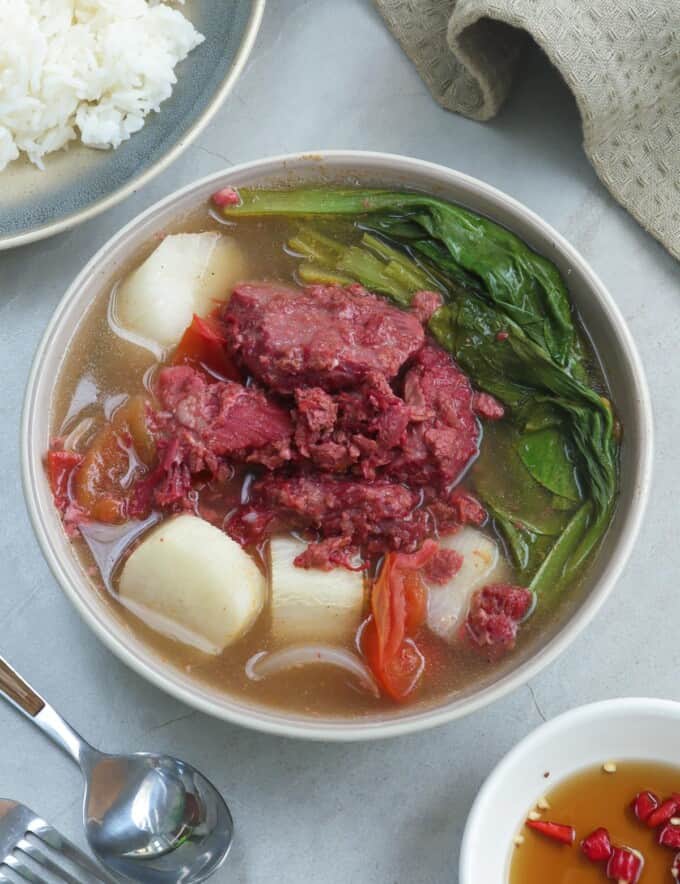 sinigang na corned beef in a white bowl with a plate of rice on the side